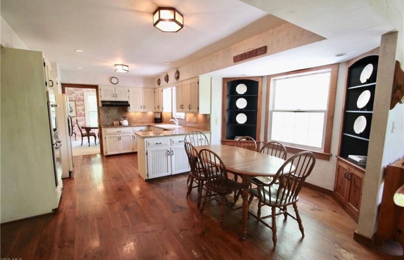 A dining area with a wooden table and chairs, adjacent to a kitchen with white cabinets and hardwood floors.
