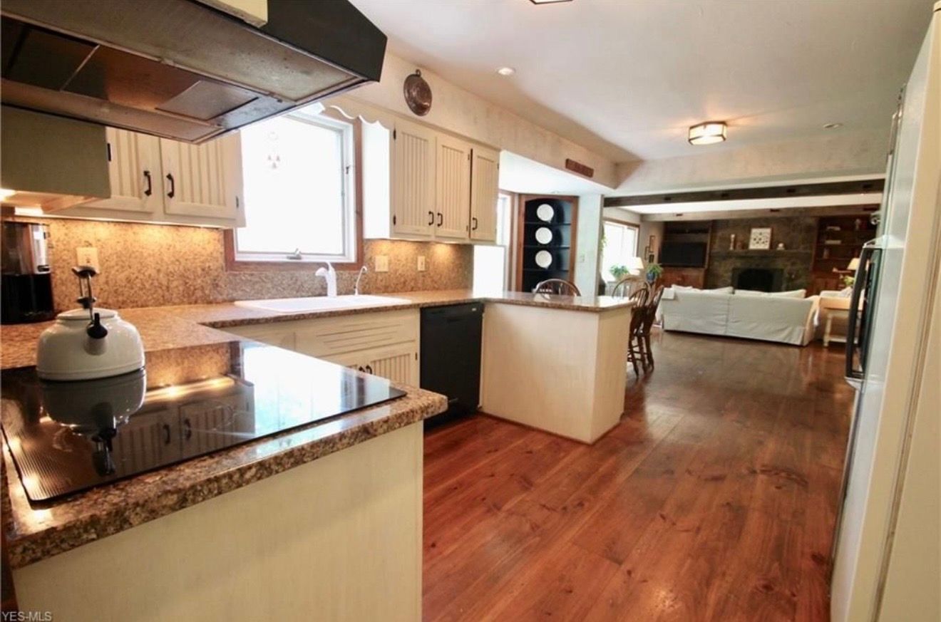 A kitchen with granite counters and white cabinets flows into a living room with wooden floors and a brick fireplace.