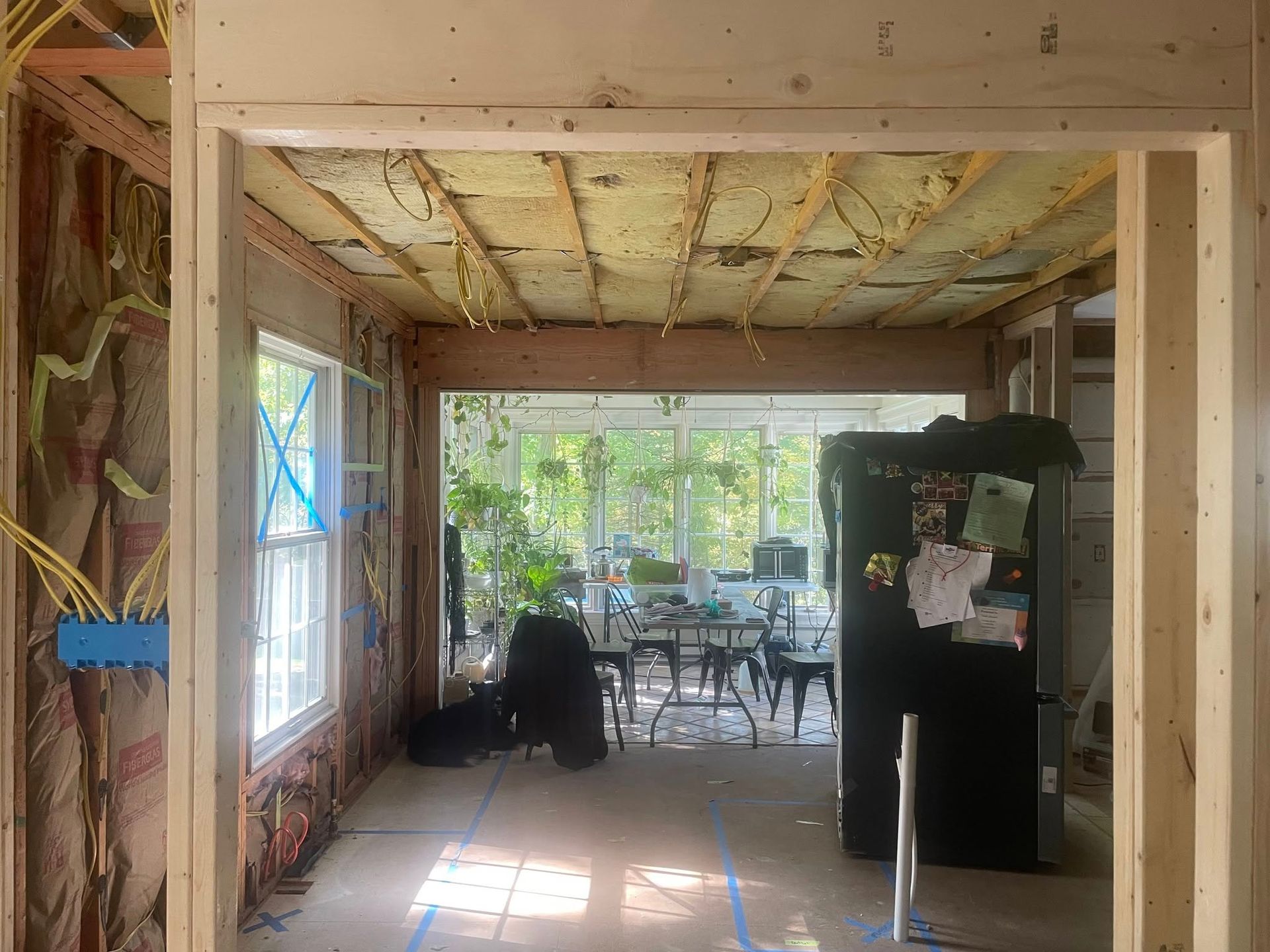 An interior view of a room under construction, showing wooden framing, exposed insulation, and a view into a sunroom.