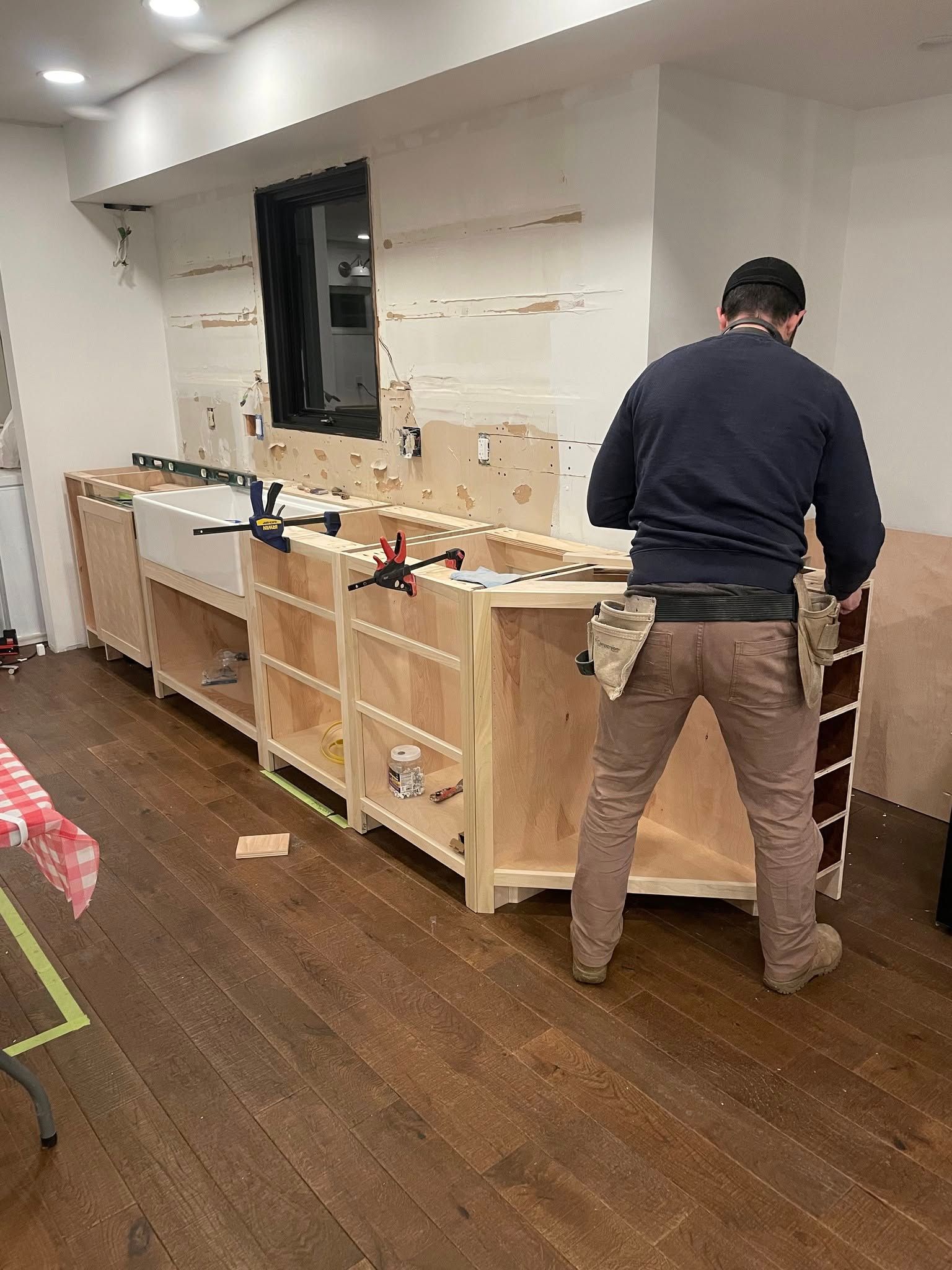 A person installs wooden kitchen cabinetry along a wall with a white farmhouse sink and wooden floors.