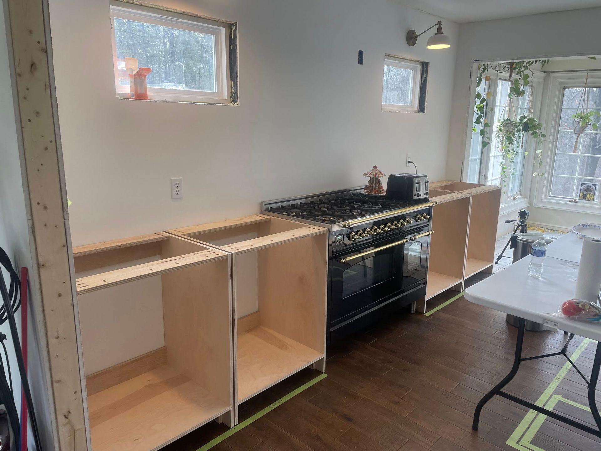Unfinished kitchen cabinets installed along a white wall next to a black stove, with windows and a folding table visible.