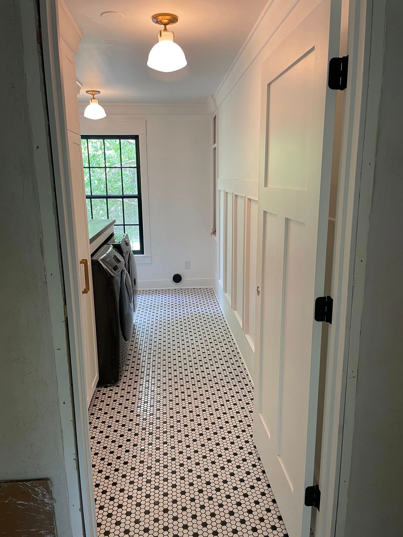 A narrow hallway laundry room with patterned black and white tile flooring, white paneled walls, and two pendant lights.