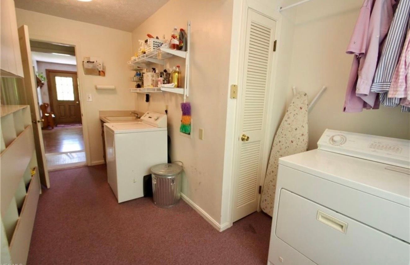 A laundry room with white washer and dryer, open wall shelving, purple carpet, and an ironing board leaning by a door.