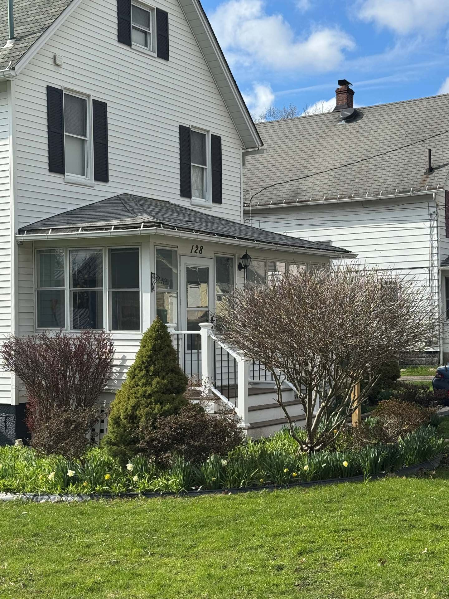 A white, two-story house with a covered front porch and green lawn, nestled next to a similar home on a sunny day.