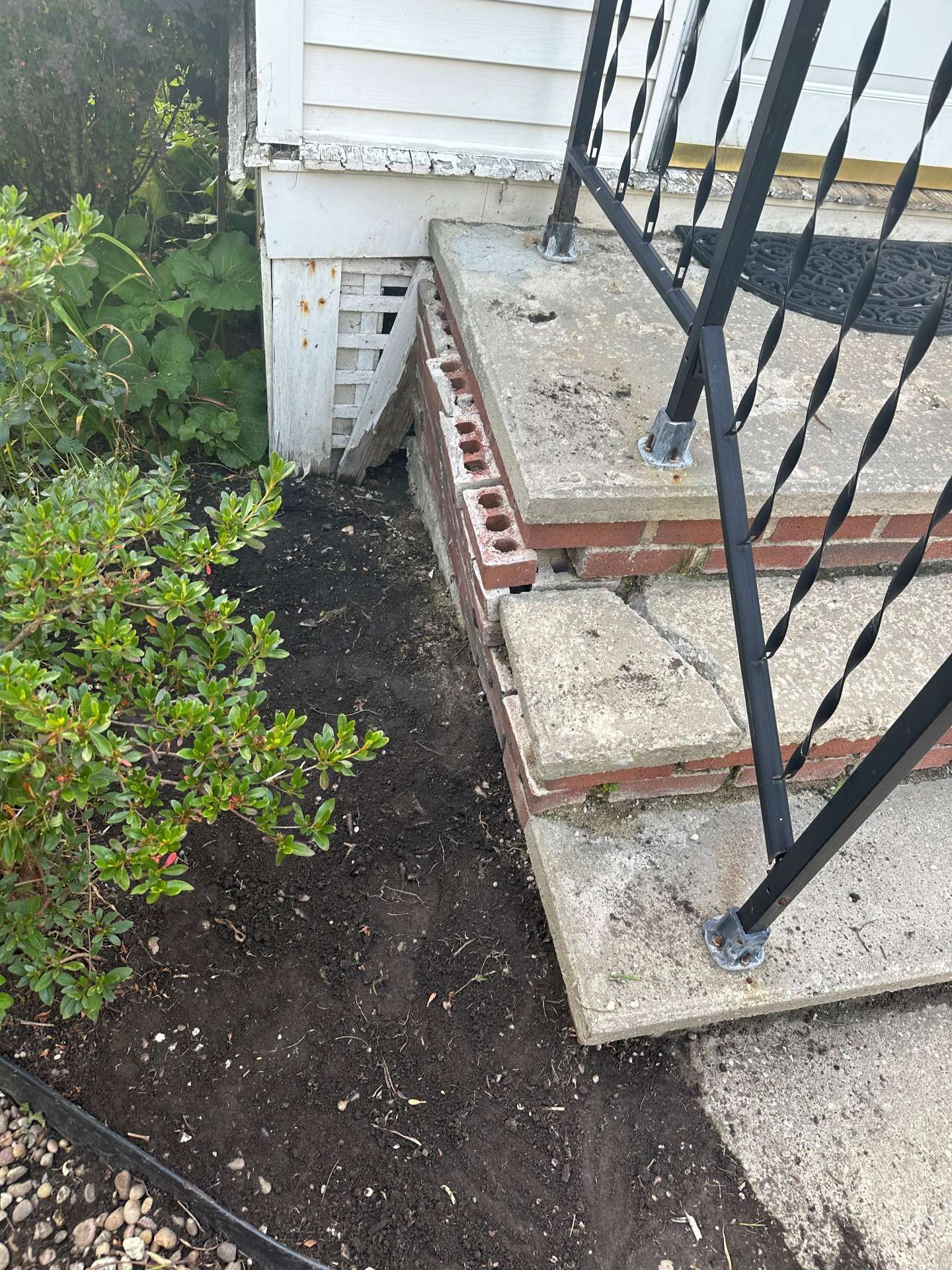 Concrete porch steps with visible gaps and crumbling brickwork meeting a house foundation, next to a garden bed.