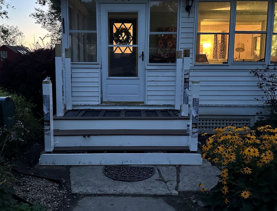White house porch steps with a storm door, illuminated by indoor lighting at dusk, next to a patch of yellow flowers.