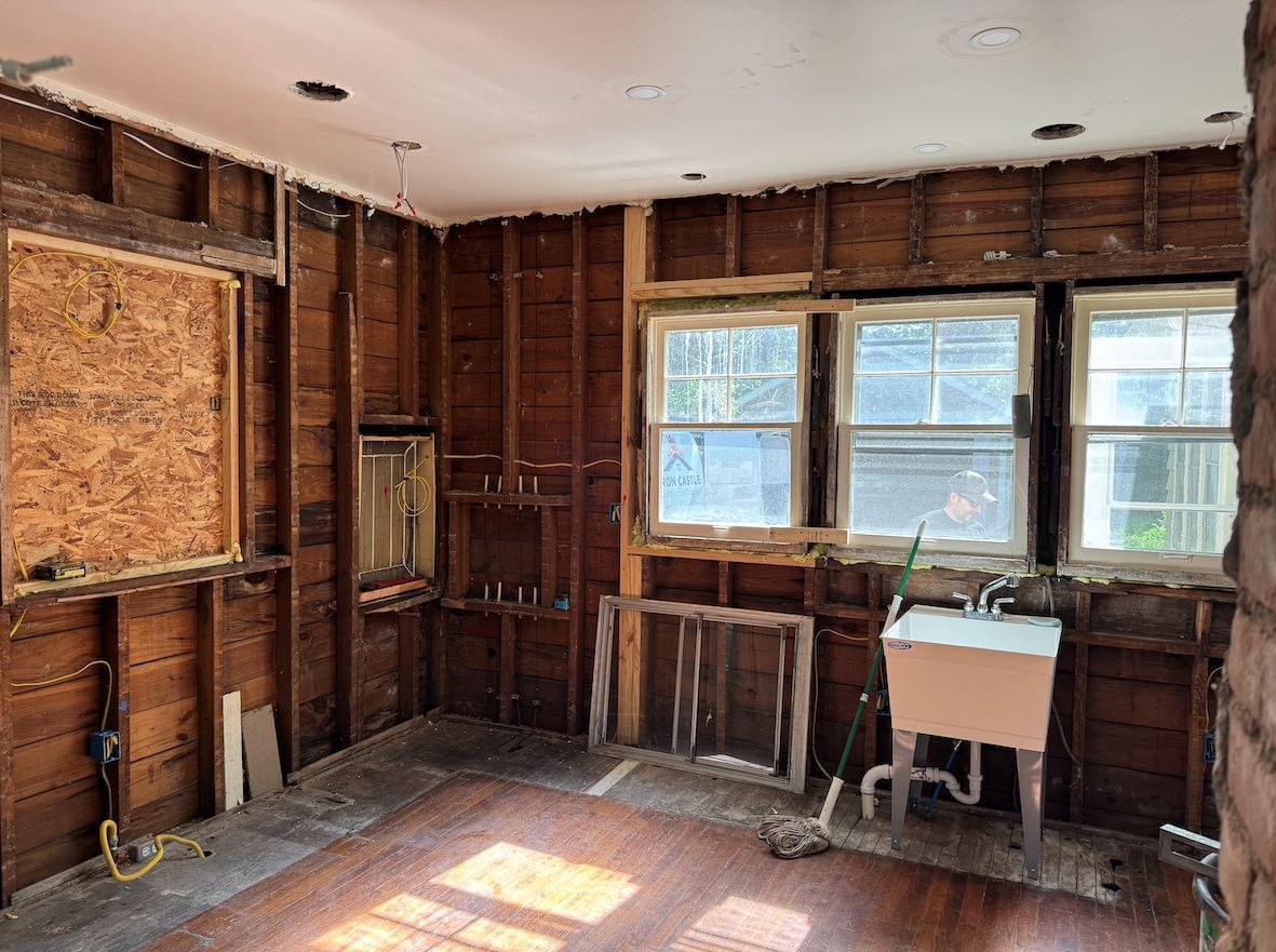 Interior view of a kitchen under renovation with exposed wood wall studs, bare windows, and a utility sink.
