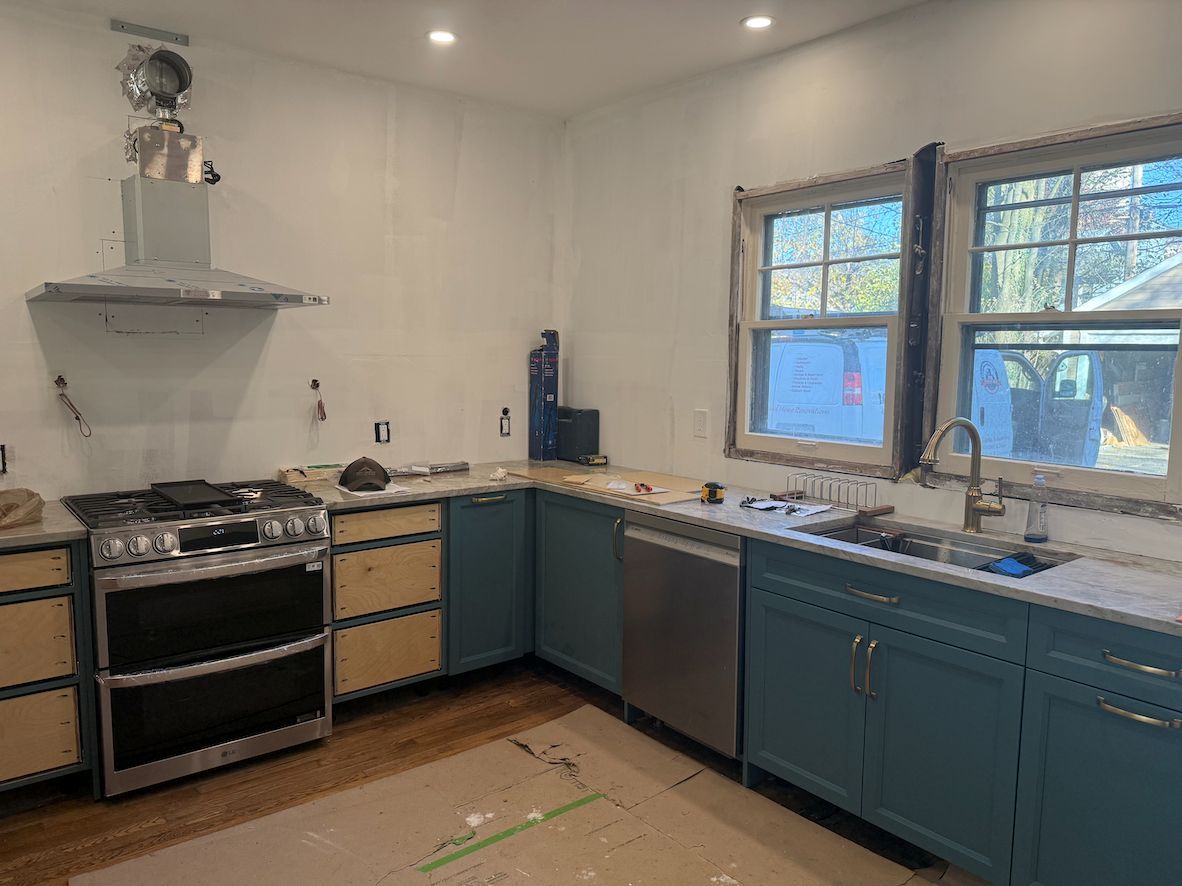 Kitchen under renovation featuring blue lower cabinets, a stainless steel stove with a vent hood, and a sink by a window.