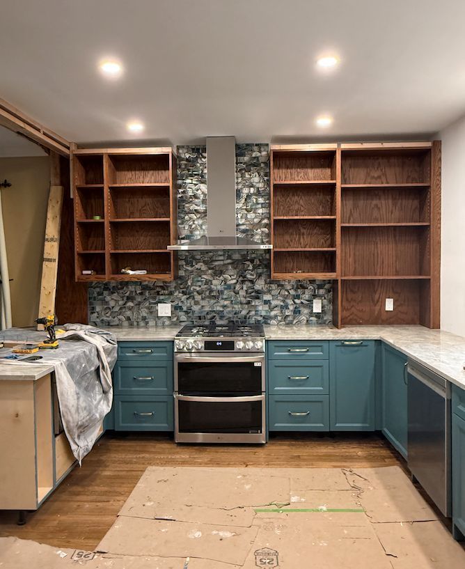 A kitchen in progress with teal base cabinets, natural wood open shelving, a stainless steel stove, and a stone backsplash.
