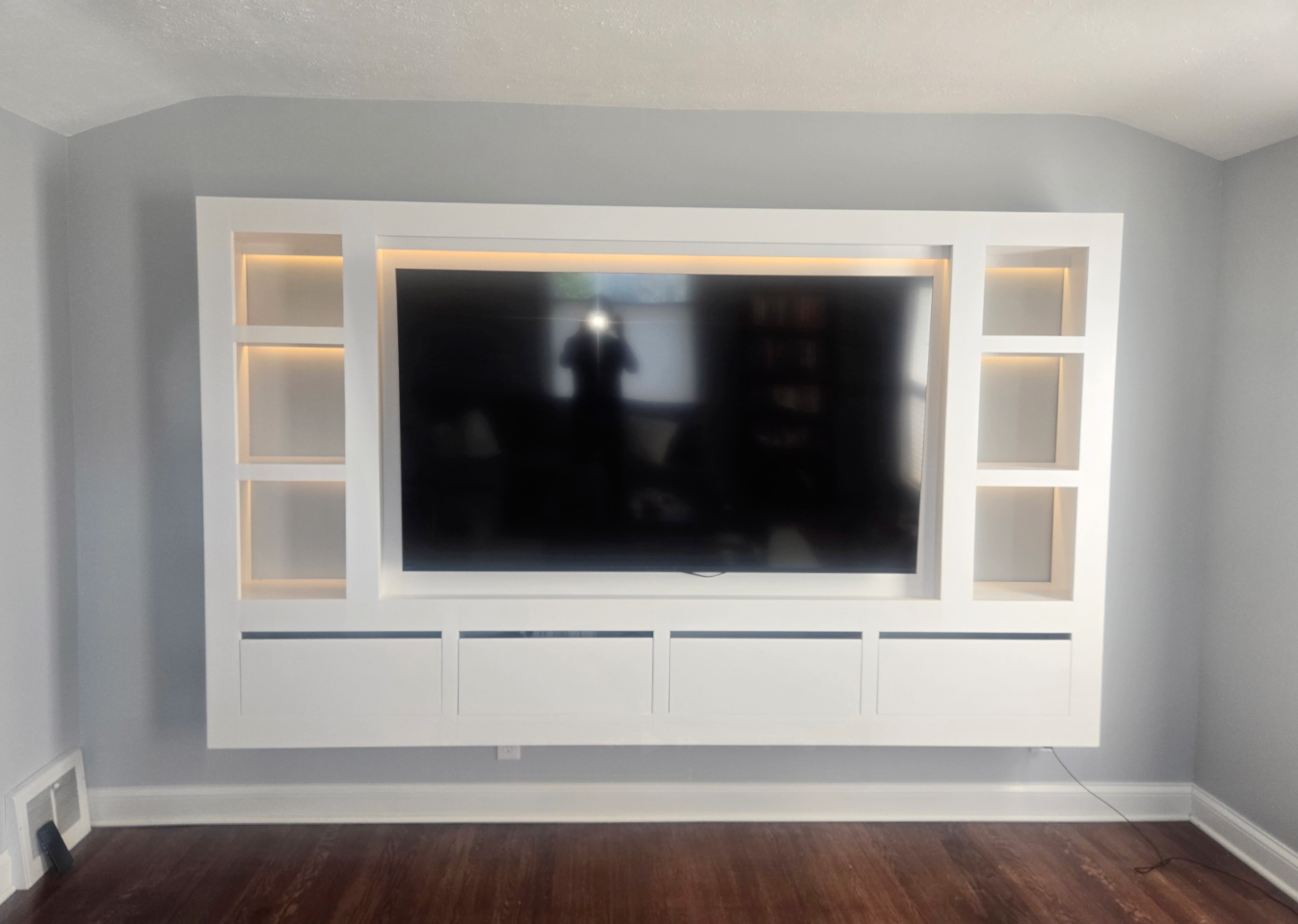 A large white built-in entertainment unit with a central television, integrated shelving, and lighting on a grey wall.