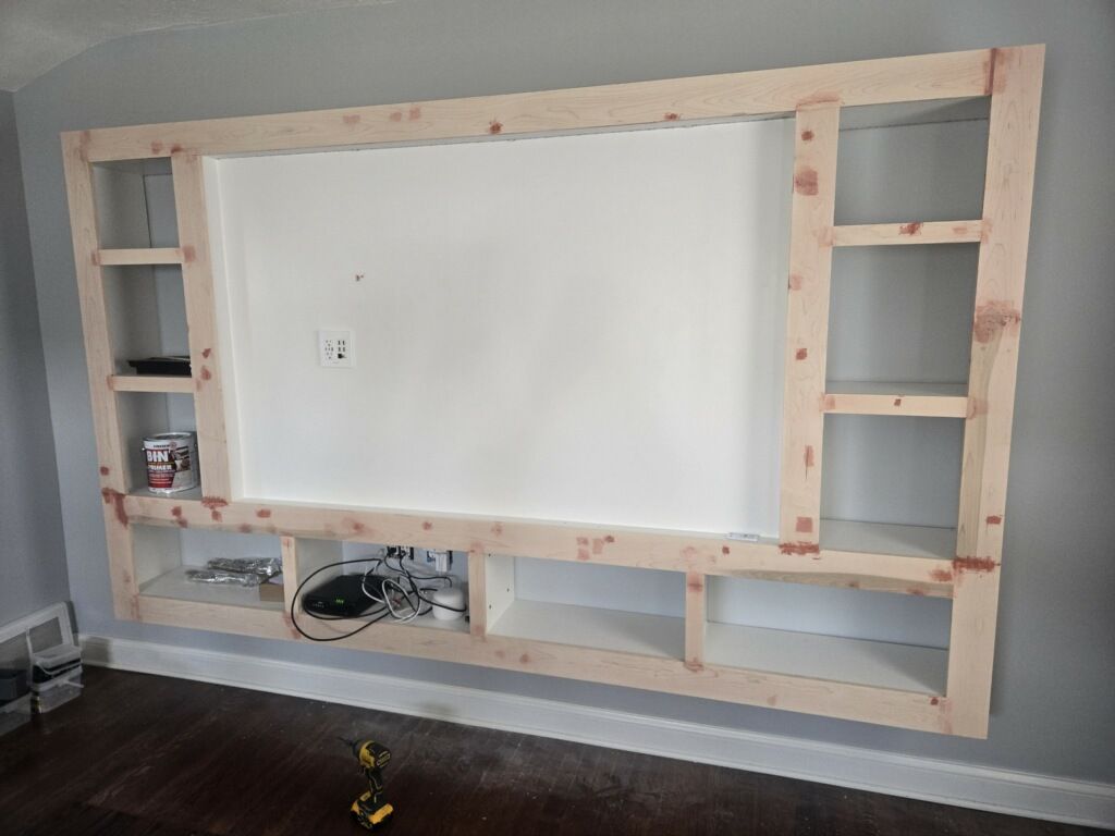A wooden entertainment center frame mounted on a gray wall with unfinished shelving and a central space for a television.