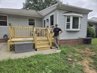 A person stands smiling beside a newly built light-colored wooden deck attached to the back of a grey house.