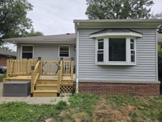 A light gray house with a brick base, a bay window, and a wooden deck with steps on the left side.