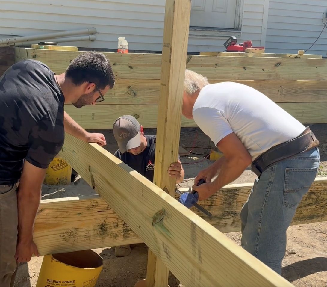 Three people work together to secure a wooden support beam onto a deck frame under construction outdoors.