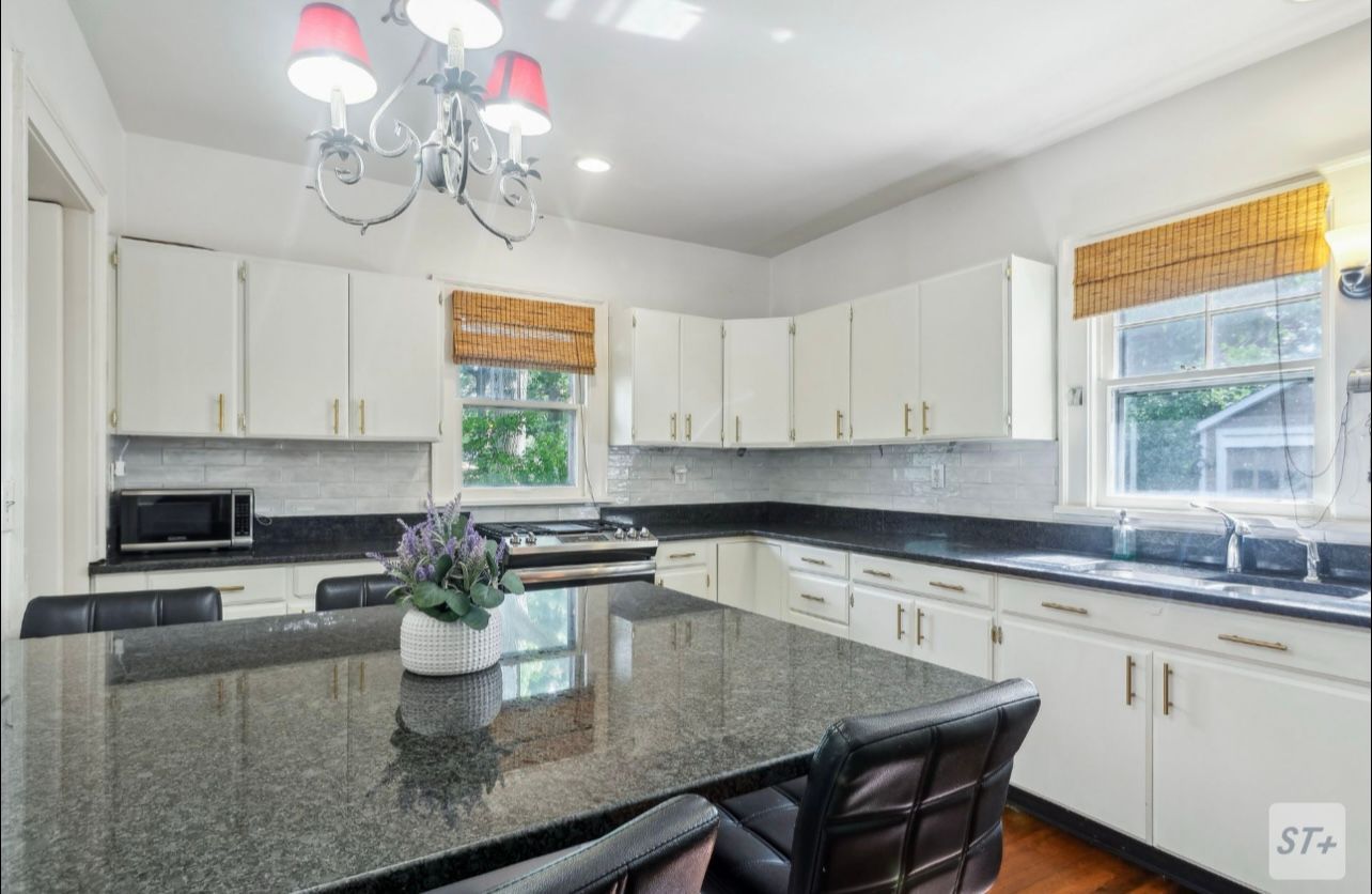 A bright, modern kitchen featuring white cabinets, a large black granite island, and a chandelier with red shades.
