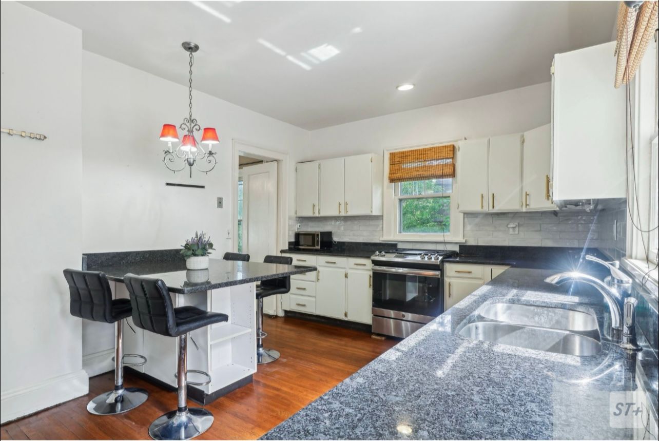 Kitchen with white cabinets, dark granite countertops, black bar stools, a small island, and a red-shaded chandelier.