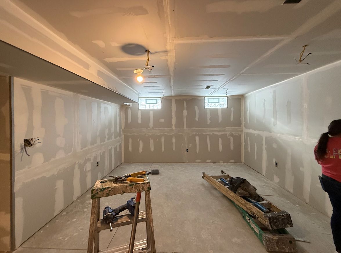 A basement under renovation with walls and ceiling covered in freshly taped drywall, a ladder, and tools on the floor.