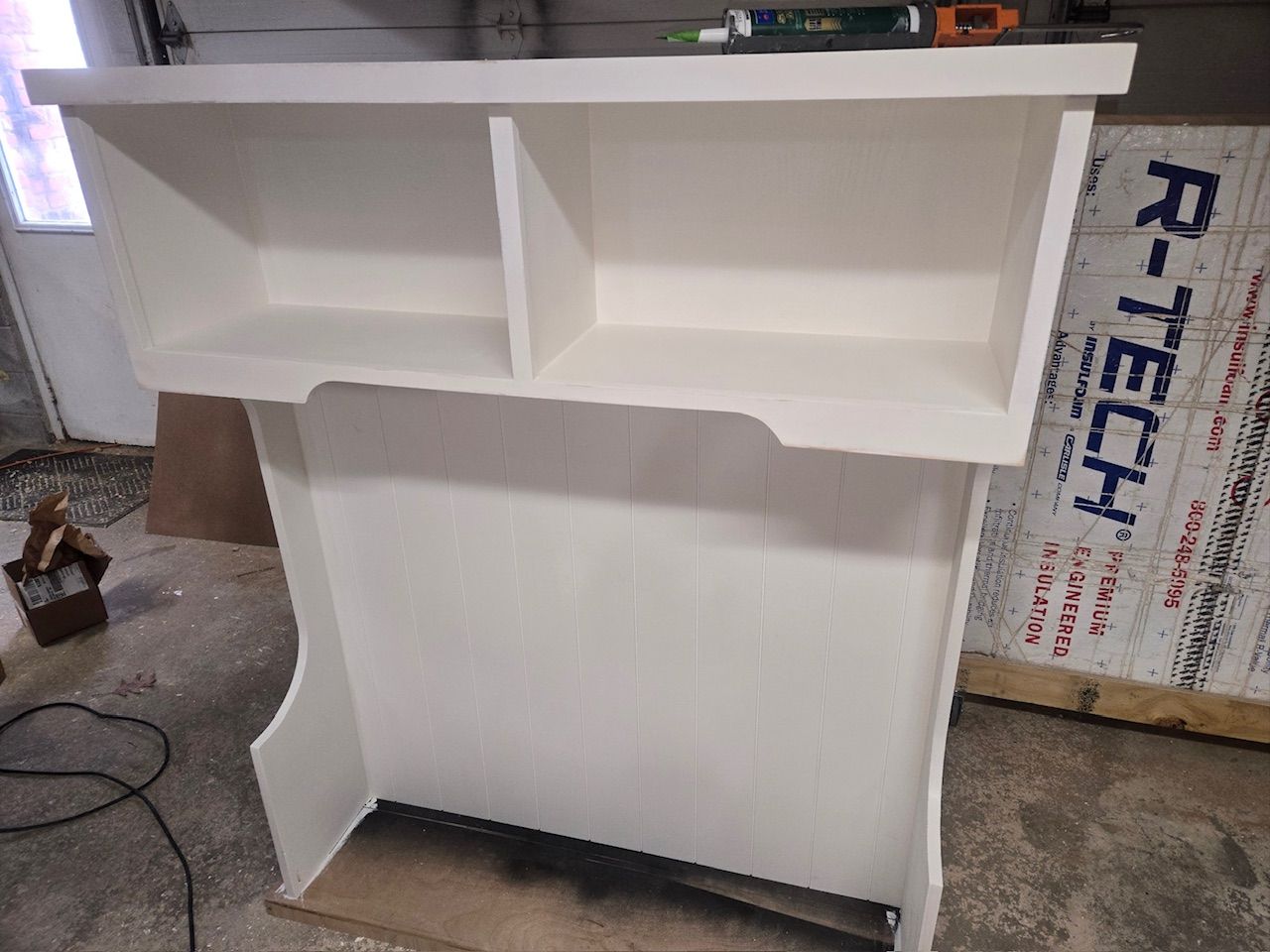 A white, unfinished wooden desk hutch with two upper compartments and curved side supports, sitting in a garage.