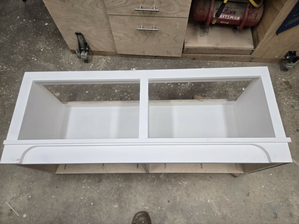 A top-down view of a rectangular, white wooden cabinet frame with two open compartments, resting on a workshop floor.