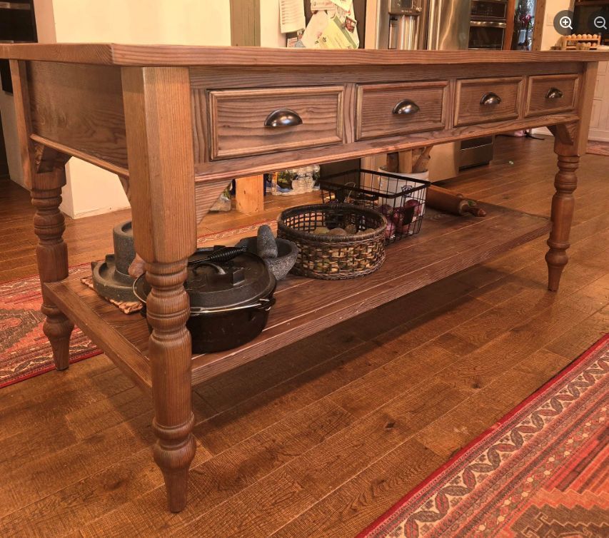 A wooden kitchen island with four drawers and a lower shelf, holding cookware and baskets on a hardwood floor.