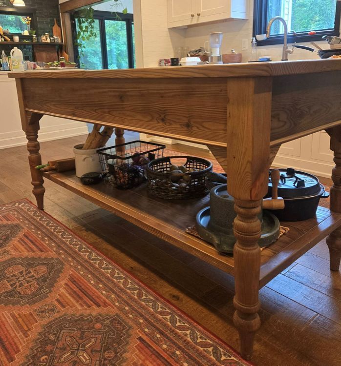 A wooden kitchen island with a lower shelf, holding baskets, cookware, and kitchen tools, placed on a patterned rug.