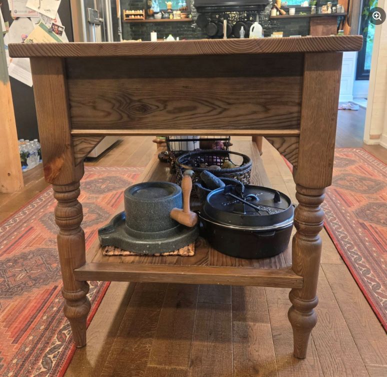 A rustic wooden kitchen island with turned legs and a bottom shelf holding a stone mortar and a black cast-iron pot.