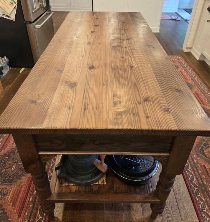 A wooden kitchen island with a natural grain finish, featuring turned legs and a bottom shelf, centered on a patterned rug.