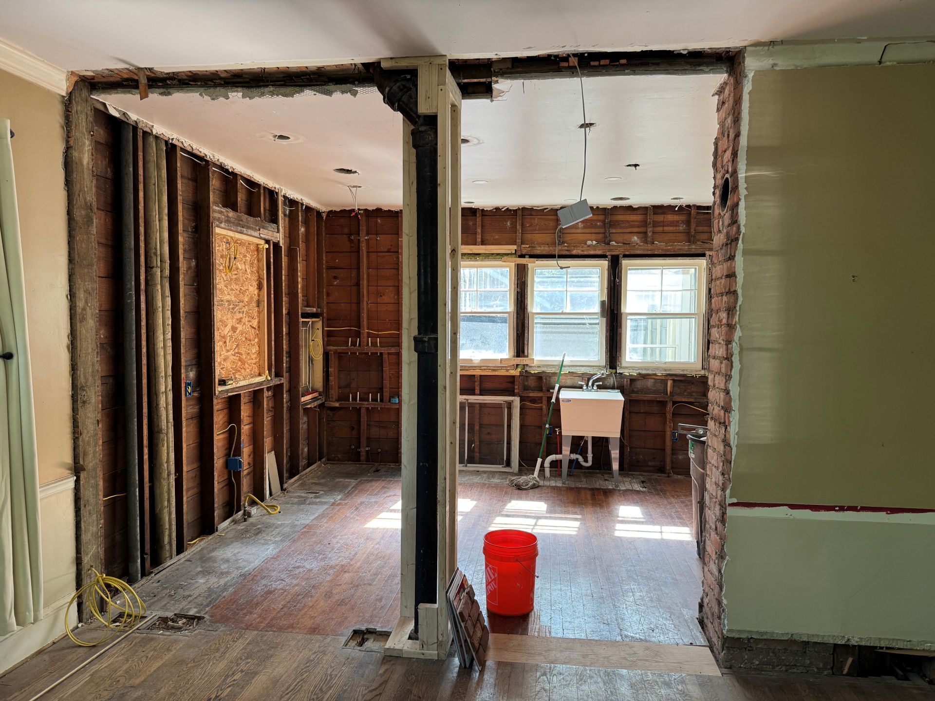 A gutted kitchen renovation with exposed wooden wall studs, a sink, and a red bucket on a hardwood floor.