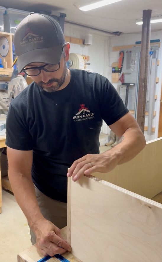 A person wearing a branded cap and shirt assembles a plywood cabinet in a workshop, carefully aligning the wood panels.