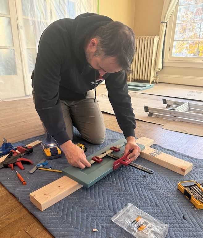 A person kneels on a drop cloth in a sunlit room, using a red hardware jig to install a hinge on a teal cabinet door.