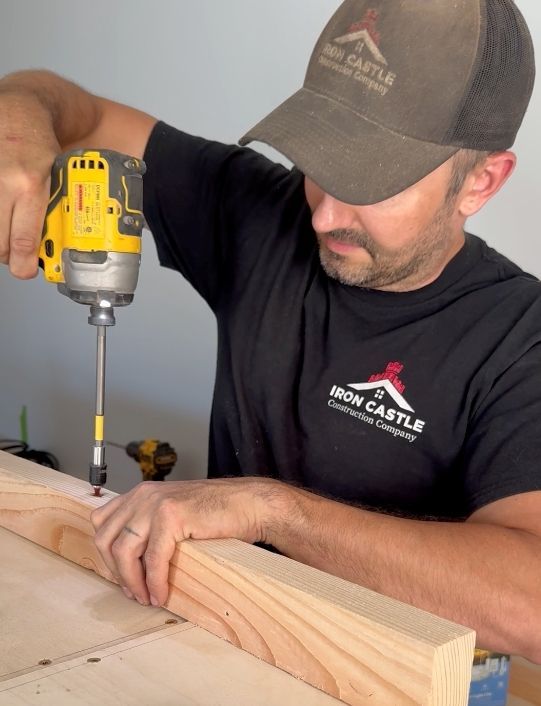 A person in an Iron Castle t-shirt and cap uses a yellow cordless drill to fasten a wooden board on a workbench.