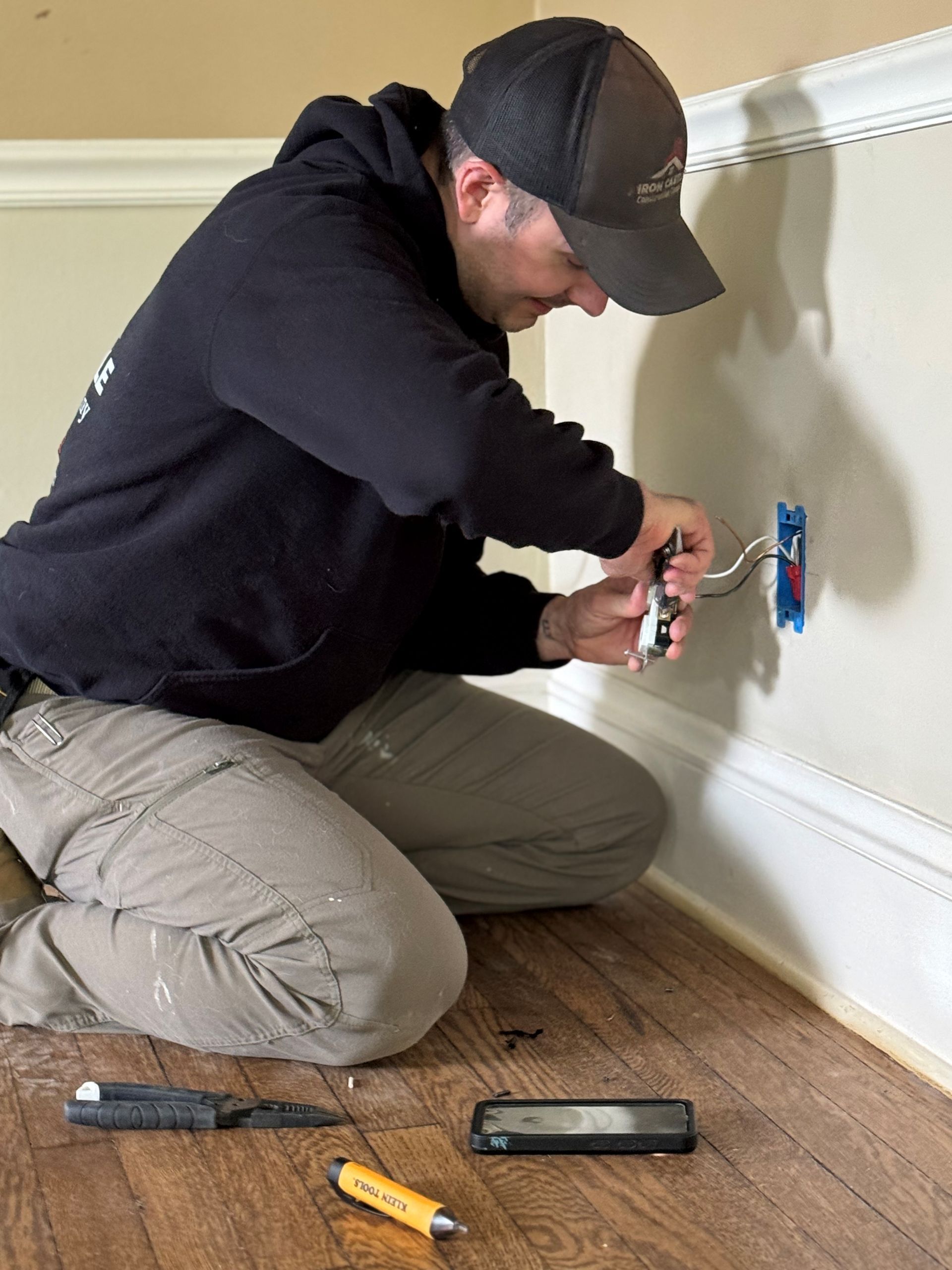 A person in a hoodie and cap kneels on a wooden floor, wiring an electrical outlet into a wall inside a room.