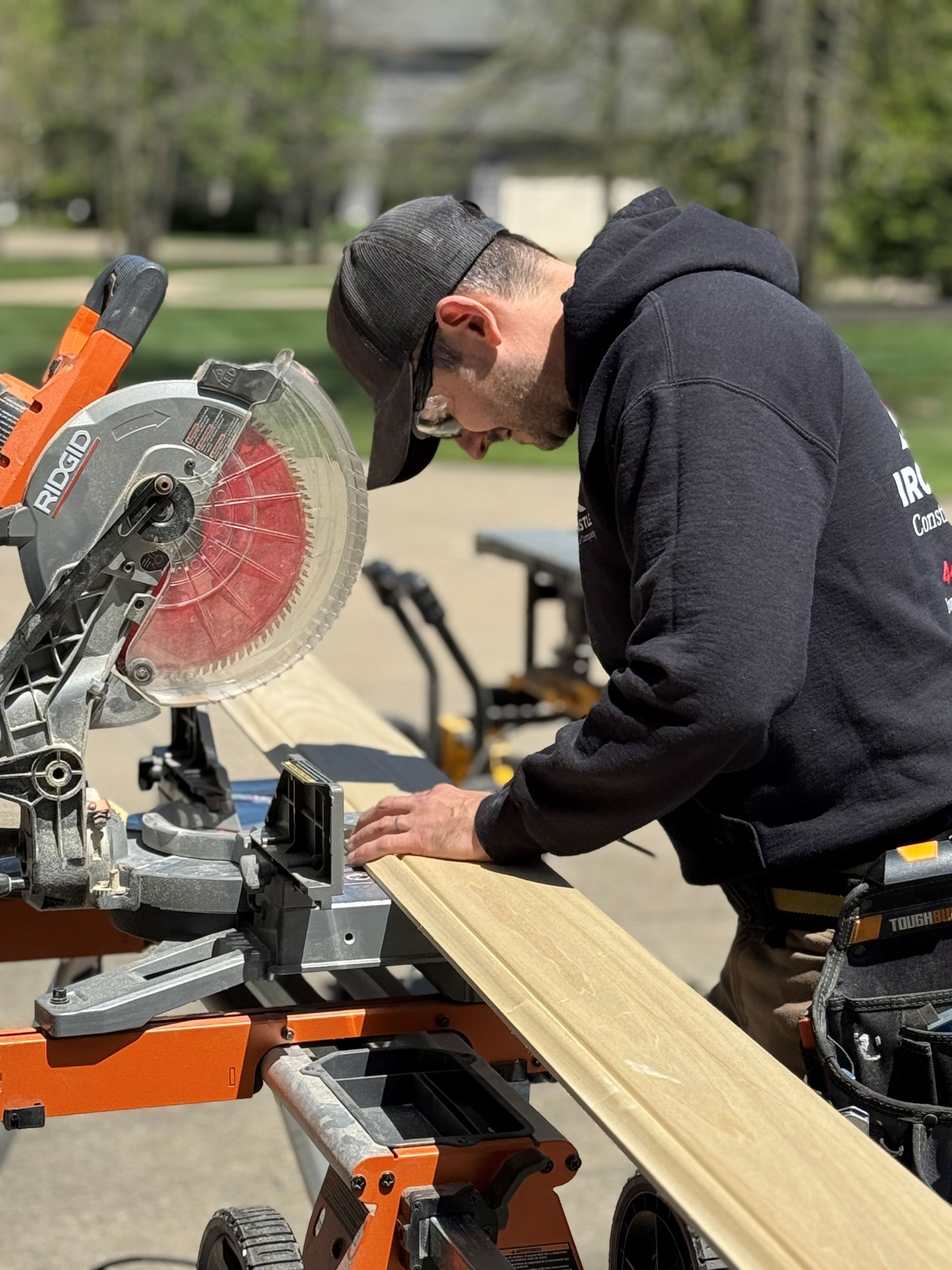 A person wearing a black hoodie and cap uses a miter saw to cut a wooden board outdoors.