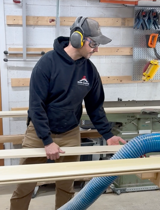 A woodworker in a workshop wears safety gear while feeding a wooden board through a jointer connected to a blue hose.