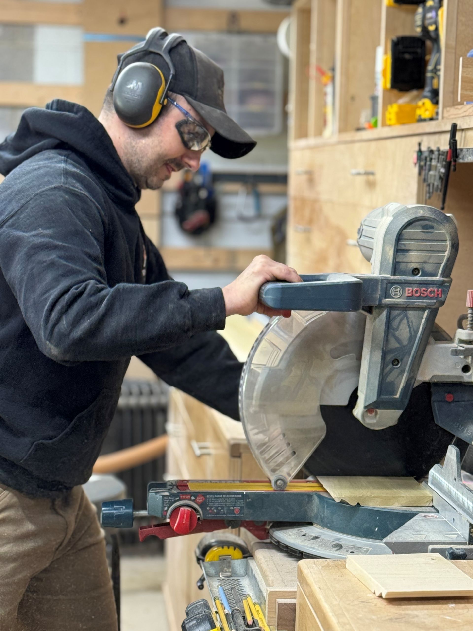 A person wearing safety glasses and earmuffs uses a miter saw to cut a piece of wood in a workshop.