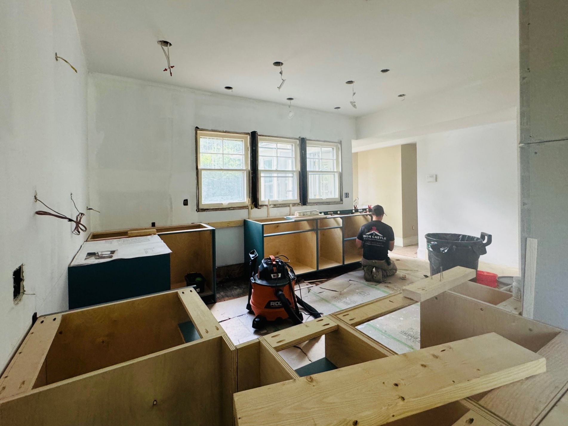 A person in a room under renovation with unfinished white walls, wooden kitchen cabinets, and construction equipment.
