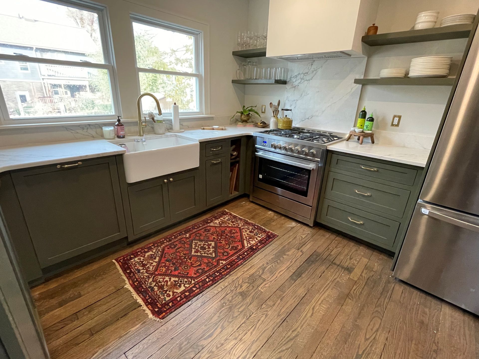 A kitchen with sage green cabinets, a white farmhouse sink, a stainless steel stove, and a red rug on hardwood floors.
