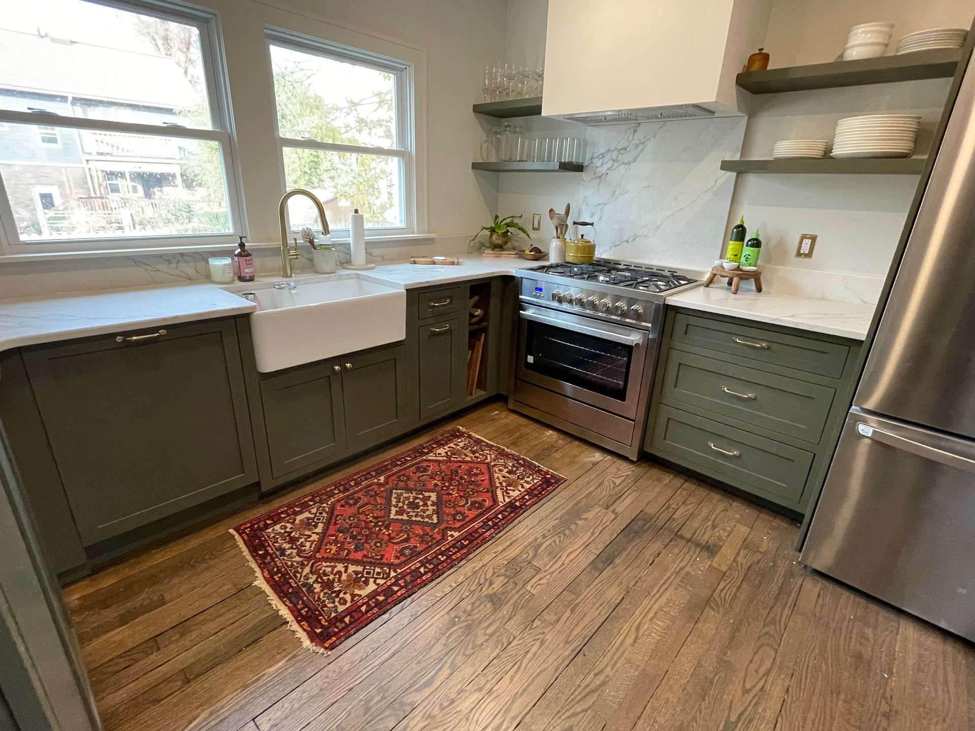 A kitchen with green cabinets, white farmhouse sink, stainless steel stove, and a red patterned rug on wooden floors.