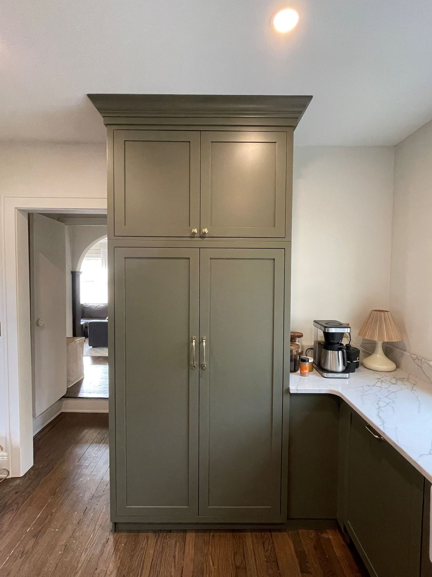 Olive-green kitchen cabinetry, featuring a tall pantry unit and corner counter space with a coffee maker and lamp.