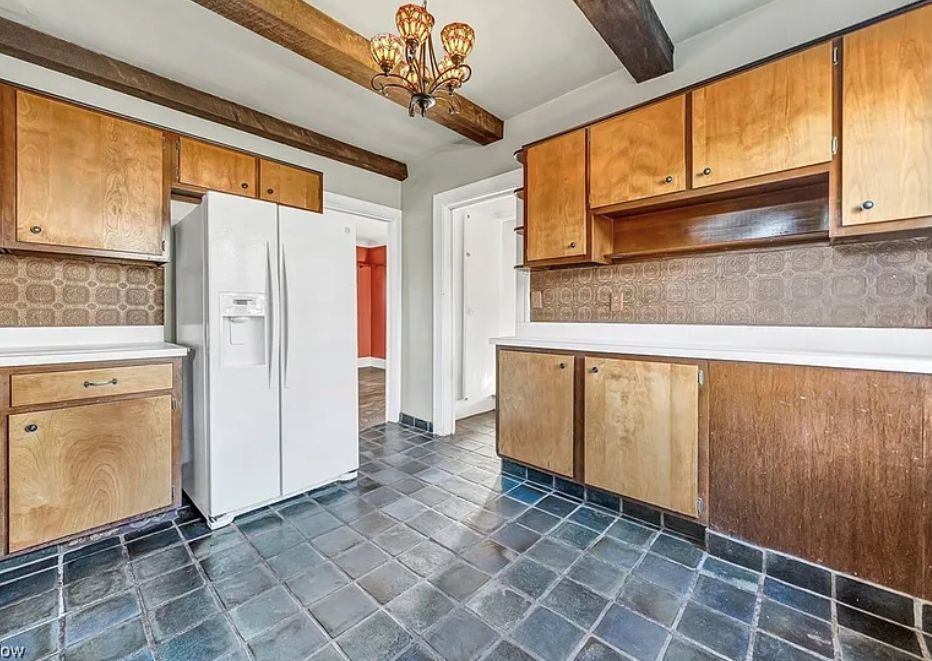 Kitchen with wooden cabinets, blue tiled floors, and a white refrigerator under a low, dark wooden beam ceiling.