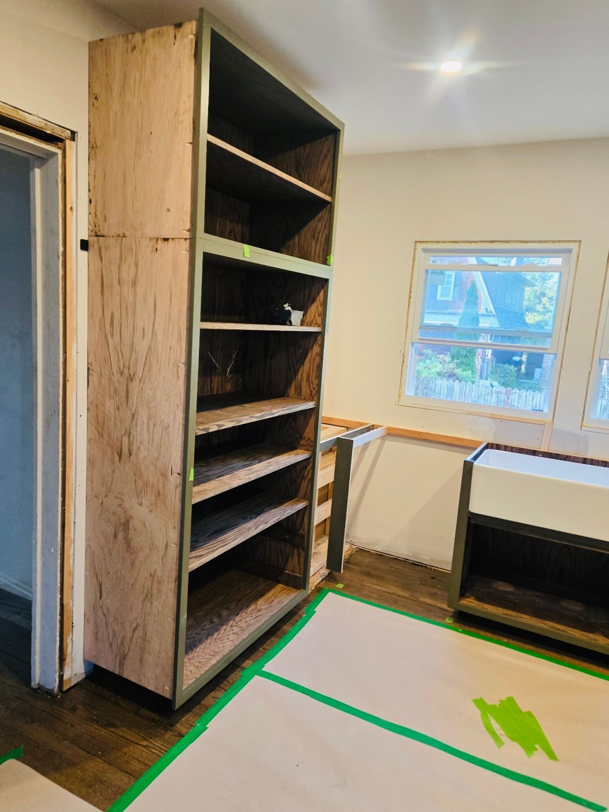Tall, unfinished wooden shelving unit next to a wall with windows and a sink in a room under construction.