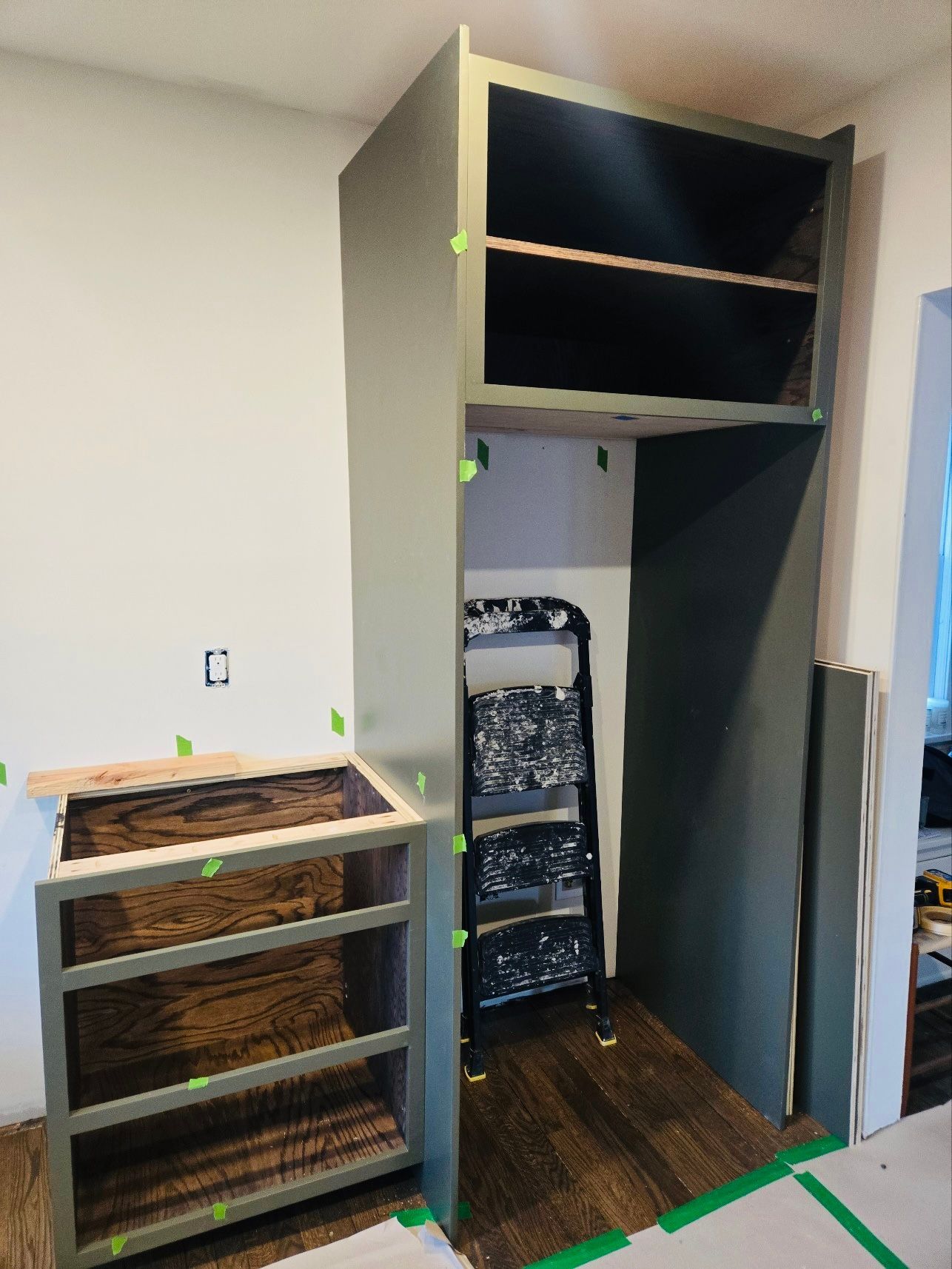 A kitchen renovation in progress, featuring a dark green cabinet frame, a partially installed base unit, and a stepladder.