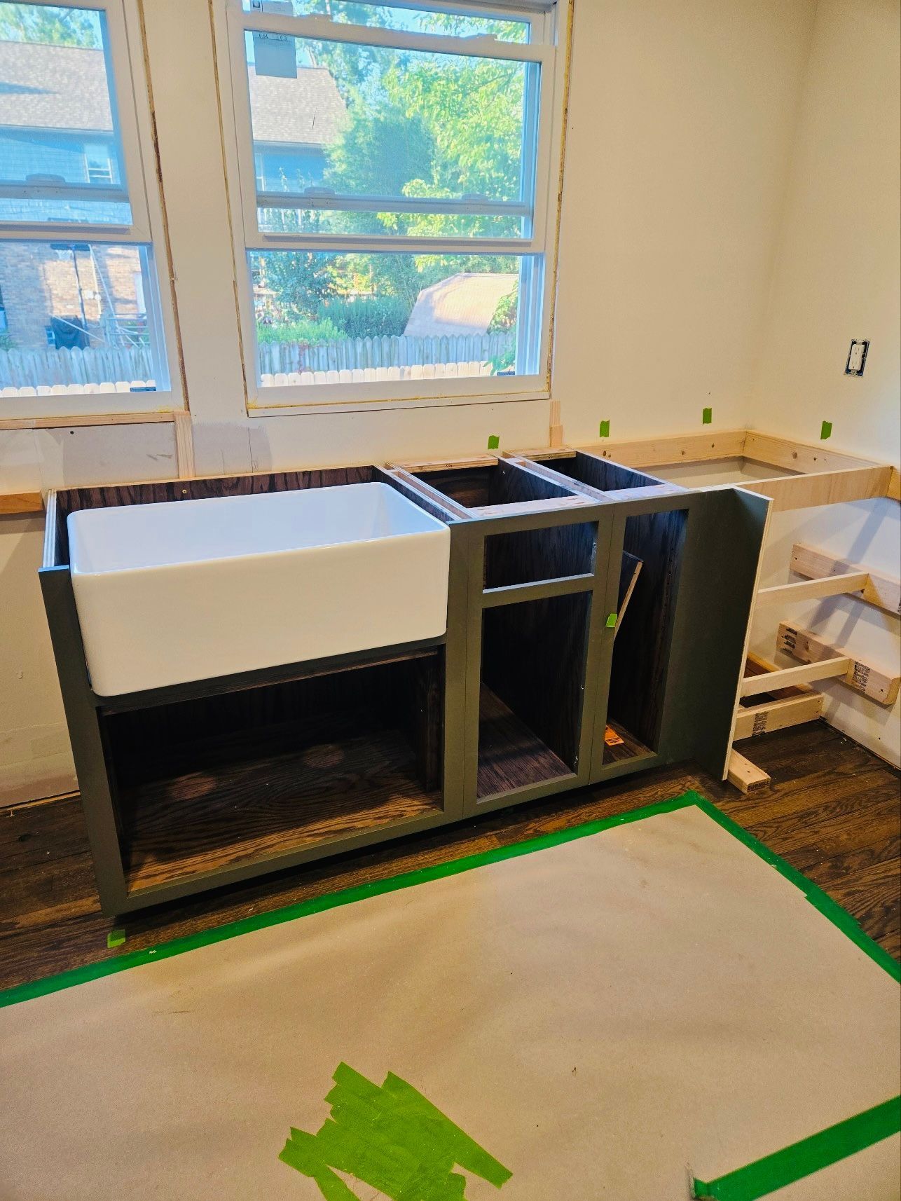 A white farmhouse sink installed in a dark grey kitchen cabinet frame under two windows, with unfinished wood framing.