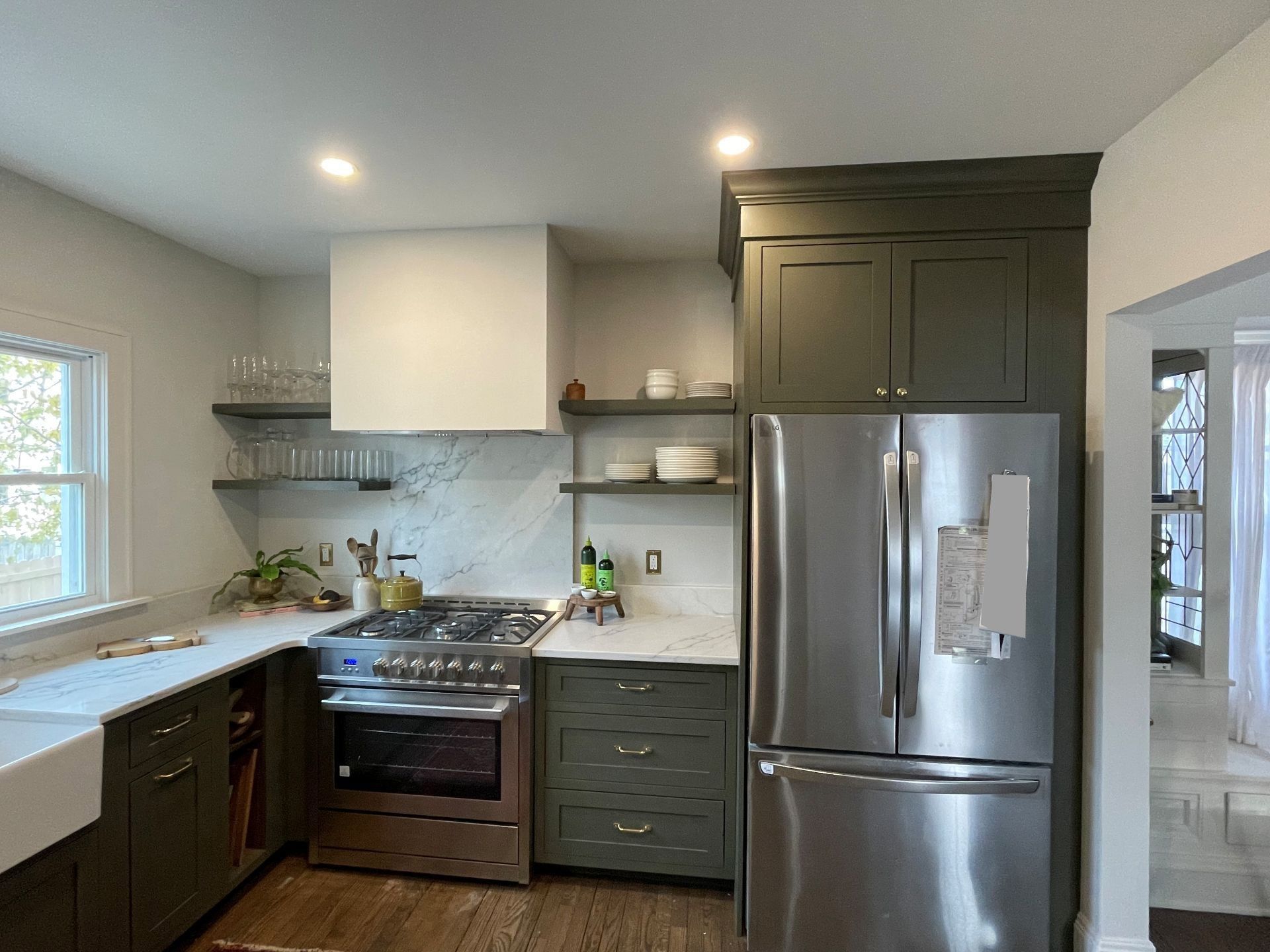 A modern kitchen with dark green cabinets, stainless steel appliances, white marble countertops, and open shelving.