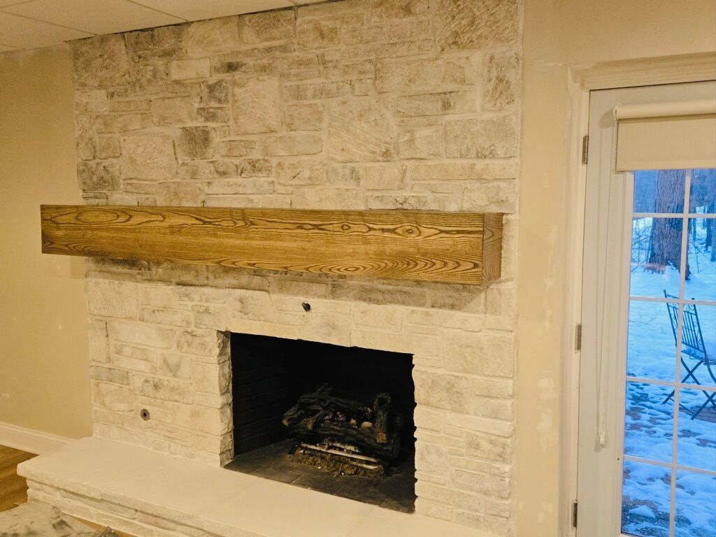 A fireplace with light-colored stacked stone, a prominent dark wood mantel, and a glass door view of snow outside.