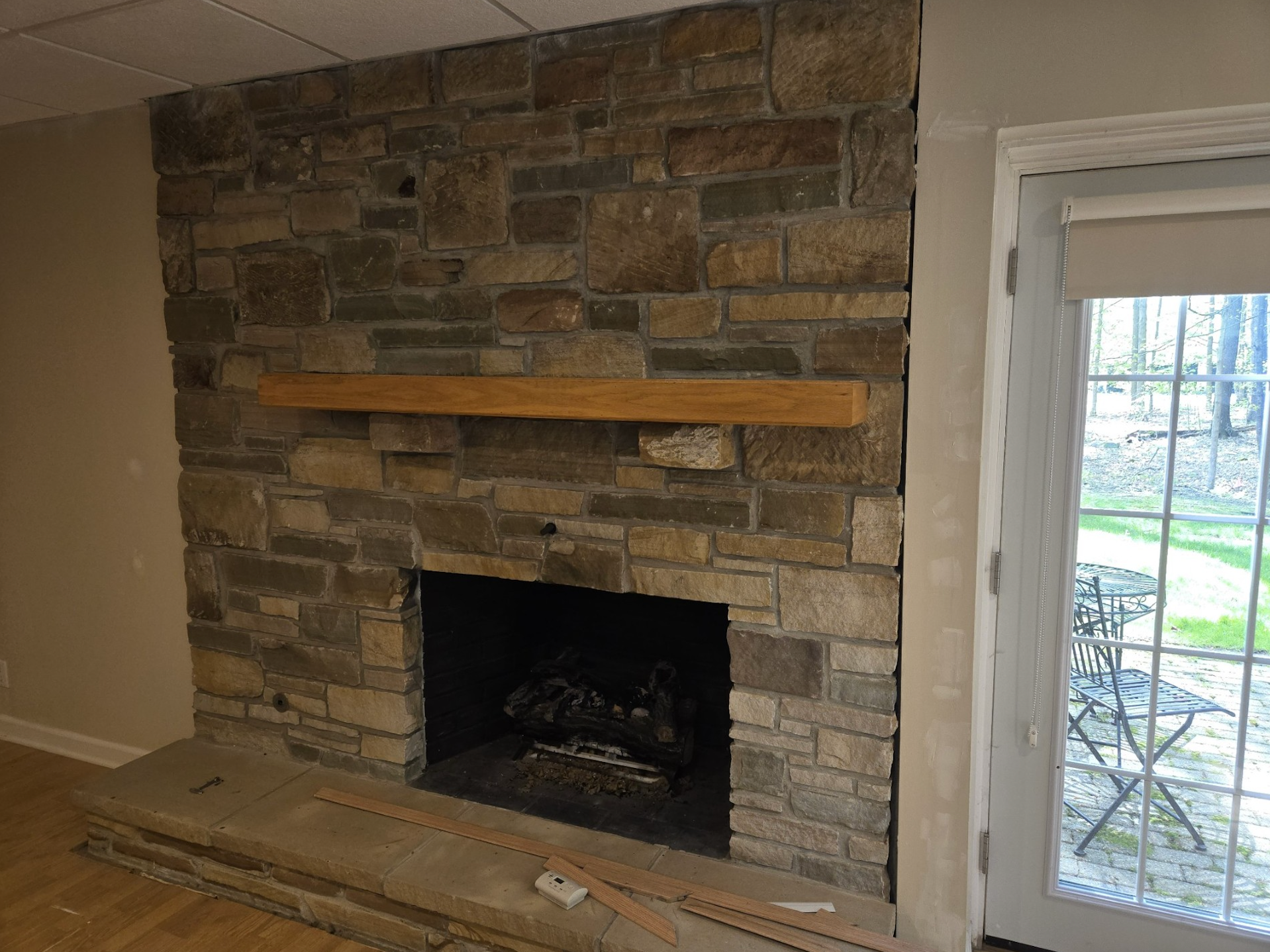 A fireplace with stacked stone masonry, a light wood mantel, a stone hearth, and a glass door leading to a yard.