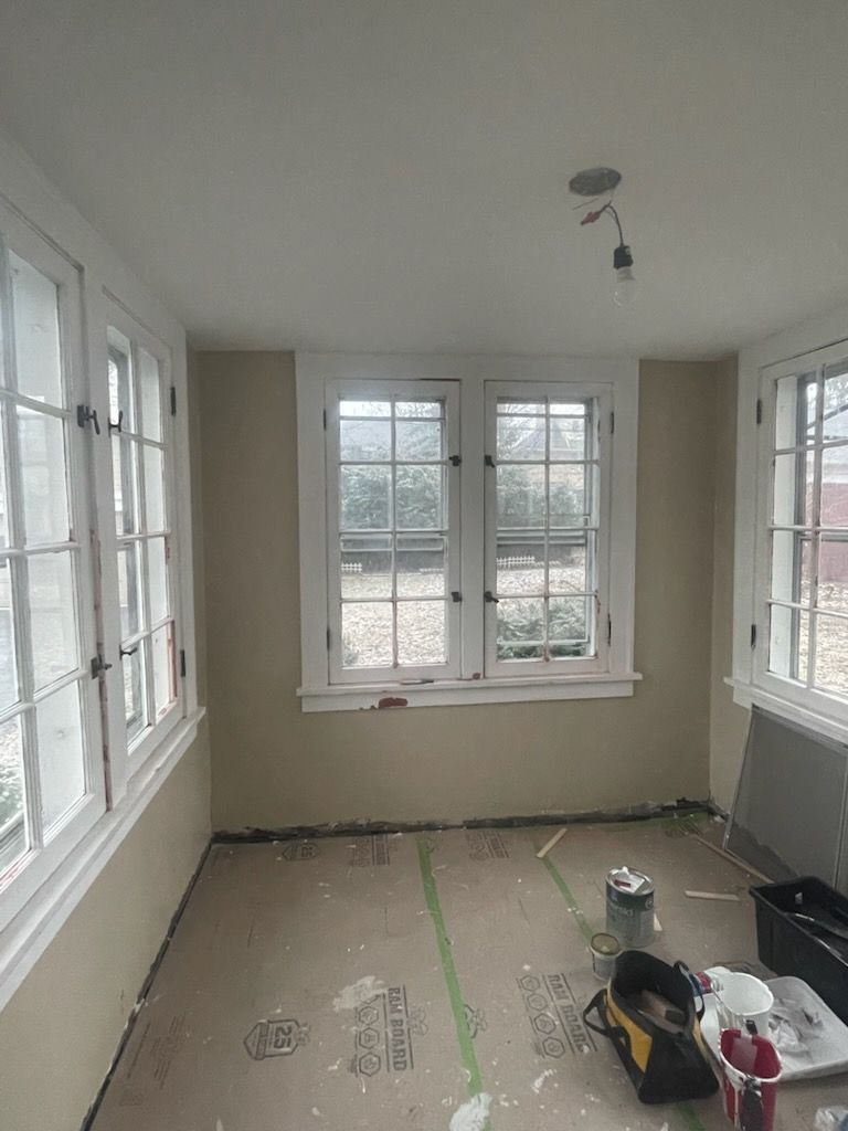A room under renovation with beige walls, white-framed windows, and unfinished flooring with construction tools.