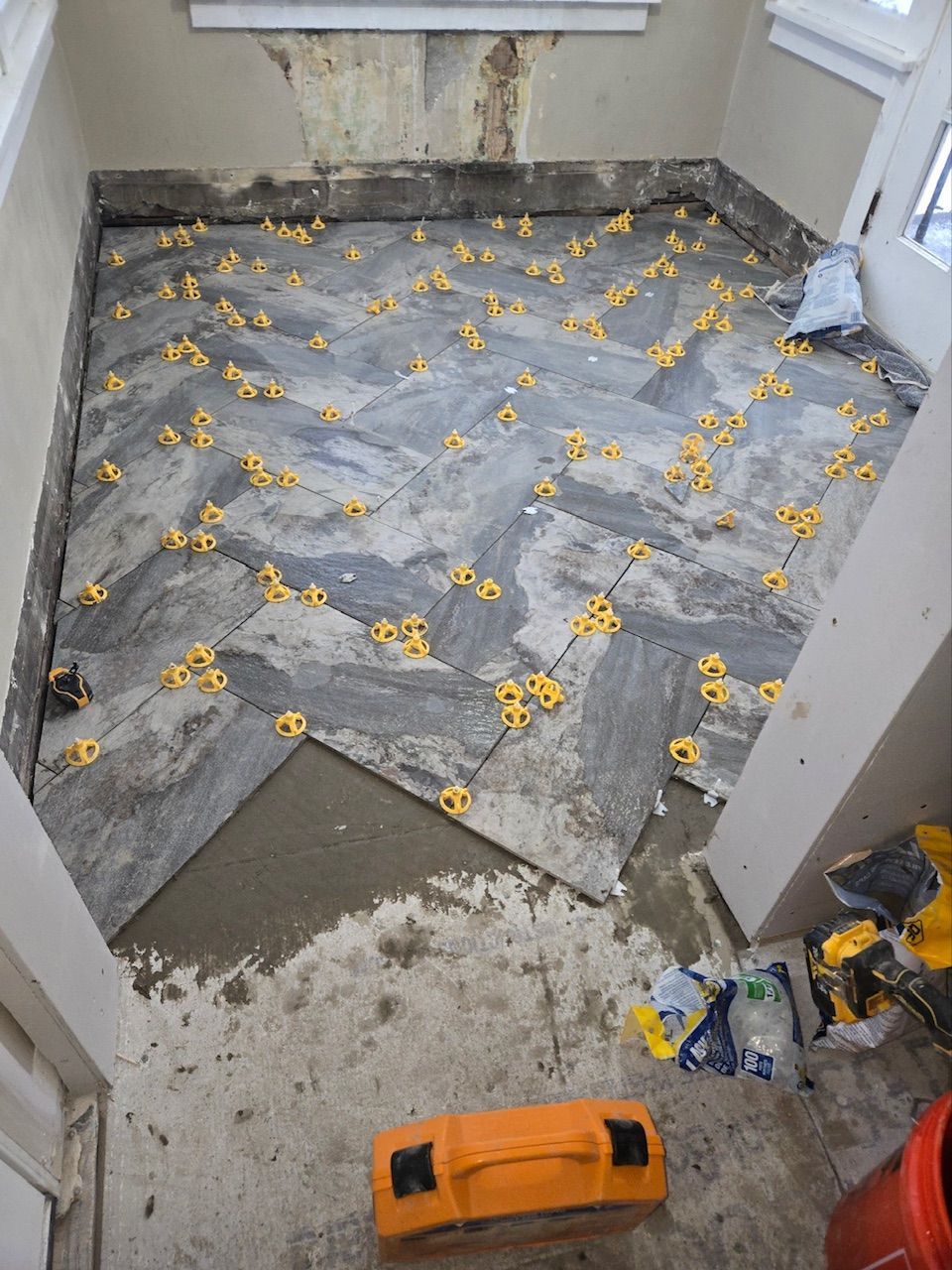 A room with unfinished floor tiling in a herringbone pattern, featuring grey wood-look tiles and yellow plastic spacers.