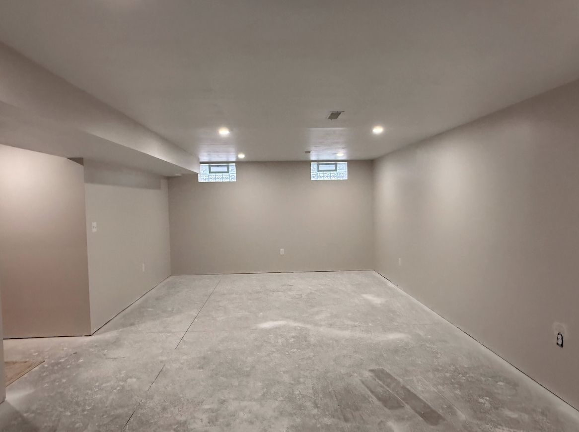 A beige, unfinished basement room with two small high-set windows, recessed lighting, and bare concrete flooring.