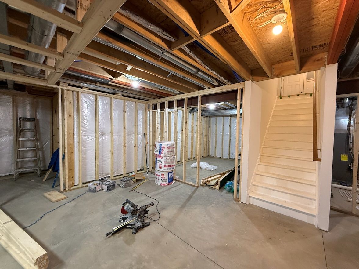 A basement under construction featuring wood framing, insulated walls, an unfinished staircase, and a power miter saw.